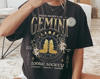 Puede incluir: Camiseta negra con un gráfico dorado del signo zodiacal Géminis. El gráfico incluye dos figuras, un círculo con símbolos astrológicos y el texto "Proud Member of Gemini Zodiac Society Intelligent Creative Charming".
