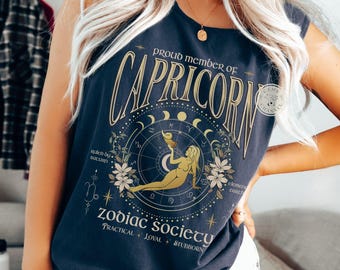 Könnte beinhalten: Marineblaues Tanktop mit goldenem Steinbock-Tierkreiszeichen-Design. Das Design enthält den Text "Proud Member of Capricorn" und "Zodiac Society" mit den Worten "Practical, Loyal, Stubborn". Die Grafik zeigt eine Frau, Blumen und astrologische Symbole.
