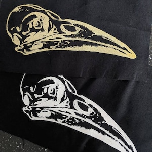 Può includere: Due toppe di tessuto nero con un design di teschio di uccello bianco e oro. Il teschio di uccello ha un lungo becco ed è rivolto a destra.