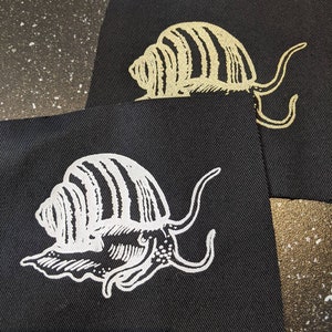 Puede incluir: Dos parches de tela negra con un diseño de caracol blanco y dorado. El caracol está estilizado con una concha y tentáculos detallados.