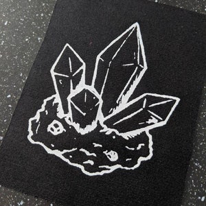 Op de afbeelding: Een zwarte canvas patch met een witte omtrek van een groep kristallen op een rots. De kristallen zijn getekend in een eenvoudige, geometrische stijl.