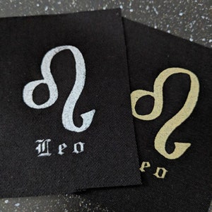 Pode incluir: Dois remendos de tecido preto com o signo astrológico Leão bordado em branco e dourado. A palavra "Leão" está escrita abaixo do signo em uma fonte gótica.