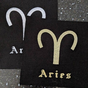 Könnte beinhalten: Zwei schwarze Stoffpatches mit dem astrologischen Symbol für Widder und dem Wort "Aries" in goldenen und silbernen Buchstaben.