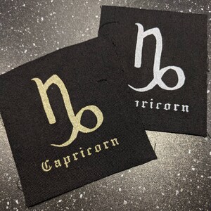 Peut inclure: Deux patchs en tissu noir avec le symbole du zodiaque Capricorne en or et en blanc. Le mot "Capricorne" est écrit sous le symbole sur chaque patch.