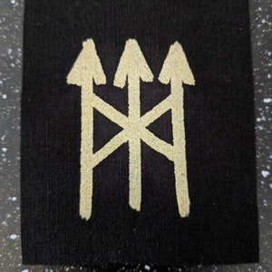 Może przedstawiać: Czarny materiałowy znaczek z białym symbolem trzech strzałek skierowanych do góry. Strzałki są połączone białym X.