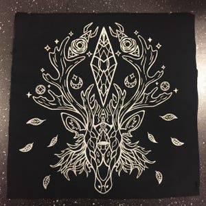 May include: A black fabric patch with a white embroidered design of a deer head with antlers. The antlers are adorned with geometric shapes and crescent moons. The deer head is surrounded by leaves and stars.