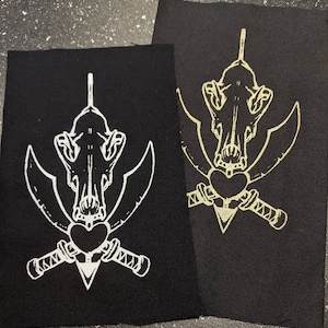 May include: Two black fabric patches with a white and gold design of a skull with crossed swords and a heart. The design is printed on the fabric.