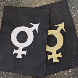 May include: Two black fabric patches with white and gold gender symbols. The symbol on the left patch is white and the symbol on the right patch is gold.