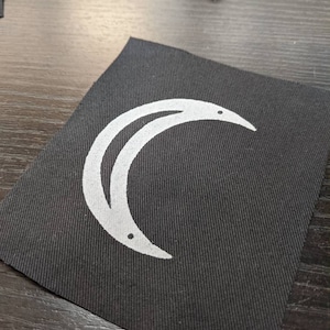 May include: A black fabric patch with a white crescent moon design. The moon has two small white dots on it.