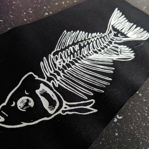 May include: A black fabric patch with a white outline of a fish skeleton. The fish is facing to the right and has a large eye.