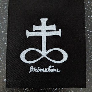 May include: Black fabric patch with a white symbol of a cross with a circle underneath. The text "Brimstone" is written below the symbol.
