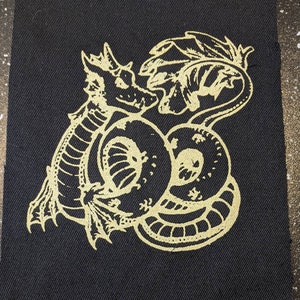 May include: A gold embroidered patch featuring a stylised dragon curled around itself on a black fabric background.