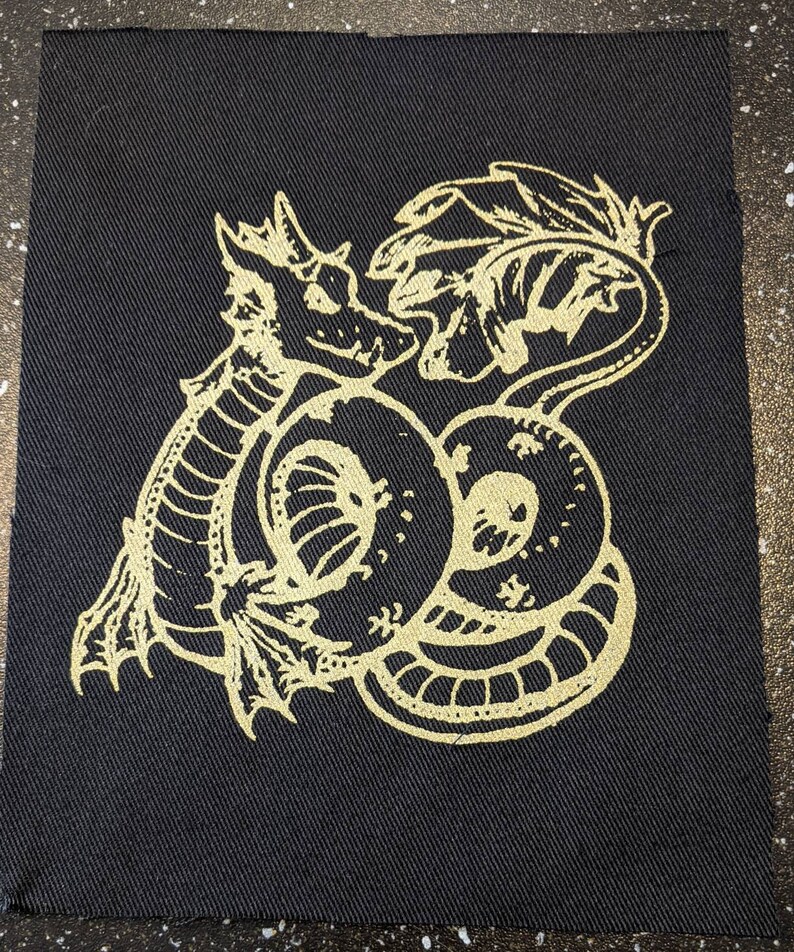 May include: A gold embroidered patch featuring a stylised dragon curled around itself on a black fabric background.