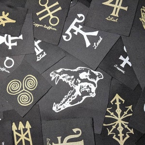 May include: A collection of black fabric patches with gold and white symbols and text. The patches feature various symbols, including a skull, a rabbit, and a cross. The text on the patches includes "Copper Saffron", "Steel", "Lead", "Arsenic", and "Vingar".