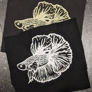 Könnte beinhalten: Zwei schwarze Stoffpatches mit weißen und goldenen gestickten Bildern von siamesischen Kampffischen. Die Fische sind nach links gerichtet und haben große, fließende Flossen.