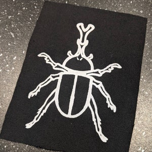 Peut inclure: Contour blanc d'un scarabée sur un patch de tissu noir. Le scarabée a une longue tête pointue et six pattes.