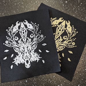 Może przedstawiać: Dwie kwadratowe czarne naszywki z motywem głowy jelenia. Jedna ma biały wzór, druga złoty. Głowa jelenia ma rogi, centralny kryształ i elementy niebieskie. Naszywki są przeznaczone do przyszywania lub naprasowywania na odzież lub akcesoria.