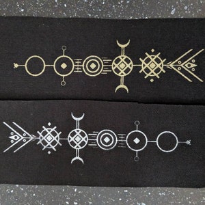 Könnte beinhalten: Zwei schwarze Stoffstücke mit einem geometrischen Muster in Gold- und Silberfaden. Das Muster ist ein symmetrisches Muster aus Kreisen, Rauten und Linien.