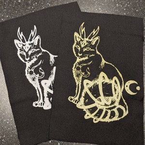 Op de afbeelding: Twee zwarte stoffen patches met een wit en gouden ontwerp van een kat met geweien. De kat heeft een derde oog en een halve maan op zijn voorhoofd. De kat zit met zijn staart om zijn lichaam gewikkeld.