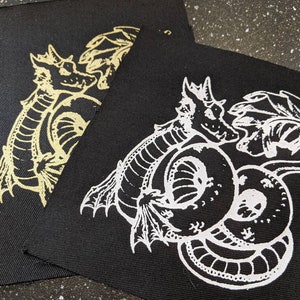 Peut inclure: Deux patchs en tissu noir avec des motifs brodés blancs d'un dragon. Le dragon est enroulé sur lui-même avec sa queue dans sa gueule. Les patchs sont de tailles différentes.