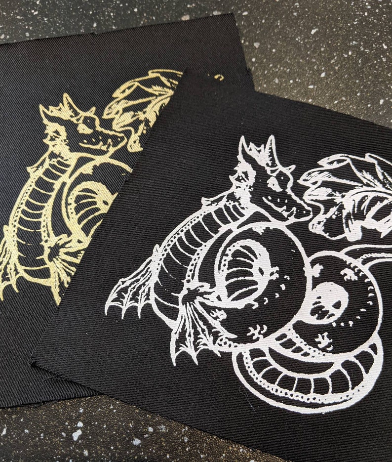 May include: Two black fabric patches with white embroidered designs of a dragon. The dragon is curled up with its tail in its mouth. The patches are different sizes.