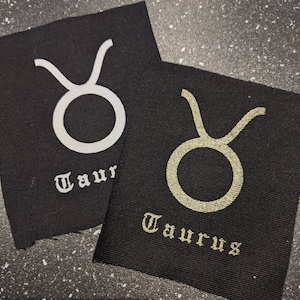 Könnte beinhalten: Zwei schwarze Stoffpatches mit dem Sternzeichen Stier in Weiß und Gold. Das Wort "Stier" ist in gotischer Schrift unter dem Zeichen geschrieben.