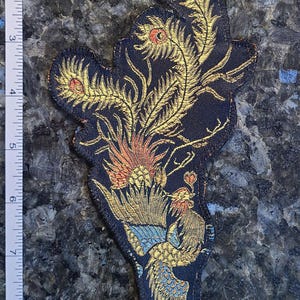 May include: A decorative patch with a detailed embroidered design of a phoenix. The patch features gold, orange, blue, and green accents on a dark blue background. The patch is approximately 4 inches in length.