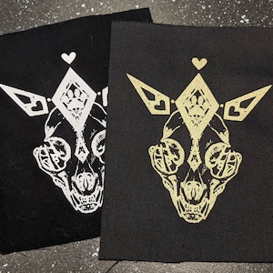 May include: Two black fabric patches with a white and gold design of a stylised skull with a heart and diamond above it. The skull has detailed eye sockets and a small heart on each side.