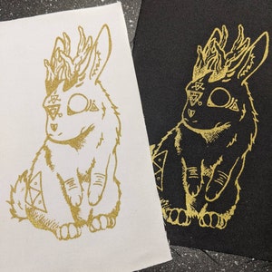 Peut inclure: Deux patchs en tissu avec un motif doré représentant un lapin avec des bois et des motifs géométriques sur son visage et son corps. Les patchs sont sur un fond blanc et noir.