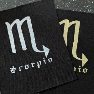 May include: Two black fabric patches with the astrological symbol for Scorpio and the word "Scorpio" in white and gold lettering.