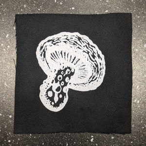 Op de afbeelding: Een zwarte stoffen patch met een geborduurd wit paddenstoel.