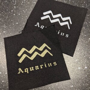 Peut inclure: Deux patchs en tissu noir avec le signe astrologique du Verseau. Le patch du haut présente le signe en blanc avec le mot "Aquarius" en blanc en dessous. Le patch du bas présente le signe en or avec le mot "Aquarius" en or en dessous.