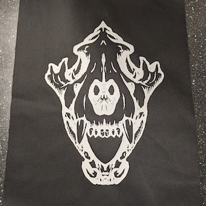 Könnte beinhalten: Eine weiße Silhouette eines Tierschädels mit offenem Maul auf einem schwarzen Stoffhintergrund.