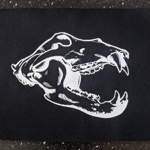 Peut inclure: Silhouette blanche d'un crâne d'animal sur un fond de tissu noir. Le crâne est tourné vers la droite et a une grande mâchoire ouverte.