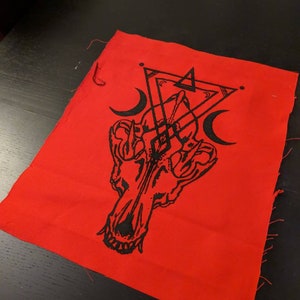 May include: Red fabric patch with a black and white illustration of a wolf skull with a geometric design above it. The design includes two crescent moons and a triangle.