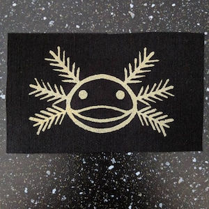 May include: A black fabric patch featuring a white axolotl design. The axolotl has a round body with two large eyes and a small mouth. It has six feathery gills on either side of its head.