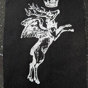 May include: Black patch with a white illustration of a winged stag wearing a crown. The stag is shown in a leaping pose, with detailed wings and antlers. The design is a stylized, monochrome image.