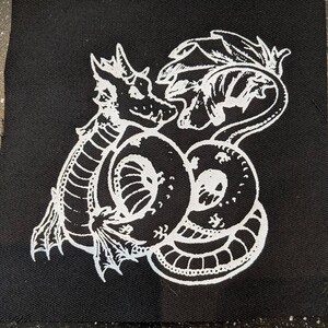 May include: White embroidered design of a dragon curled up on a black fabric patch. The dragon has a long tail and wings.