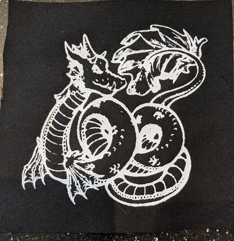 May include: White embroidered design of a dragon curled up on a black fabric patch. The dragon has a long tail and wings.