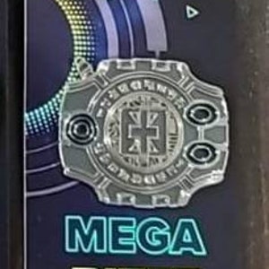 May include: A silver-colored pin with a circular design featuring a stylized cross in the center. The pin has a black background with a blue and green gradient. The text "MEGA BITEZ" is printed in green and blue letters below the pin.