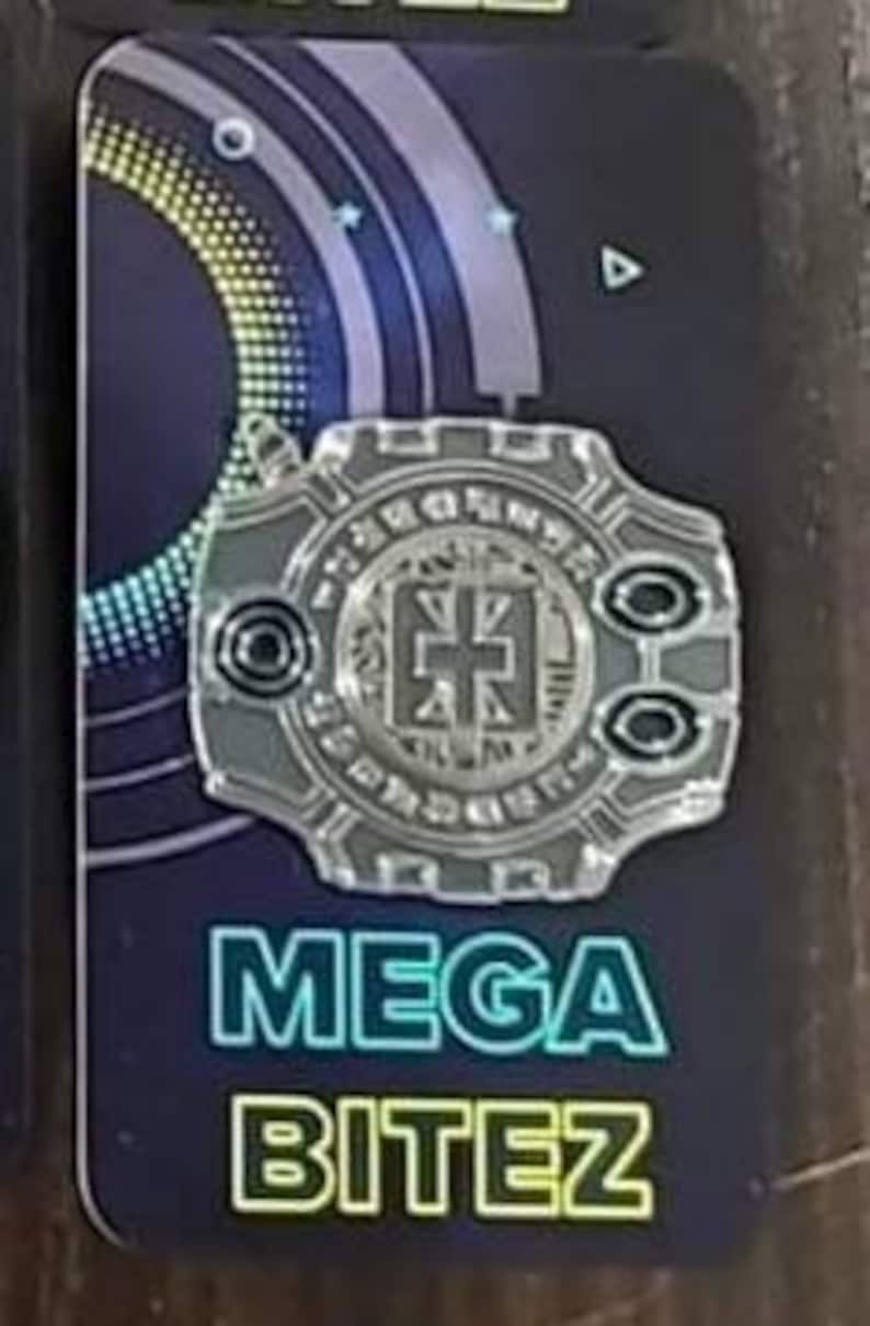 May include: A silver-colored pin with a circular design featuring a stylized cross in the center. The pin has a black background with a blue and green gradient. The text "MEGA BITEZ" is printed in green and blue letters below the pin.