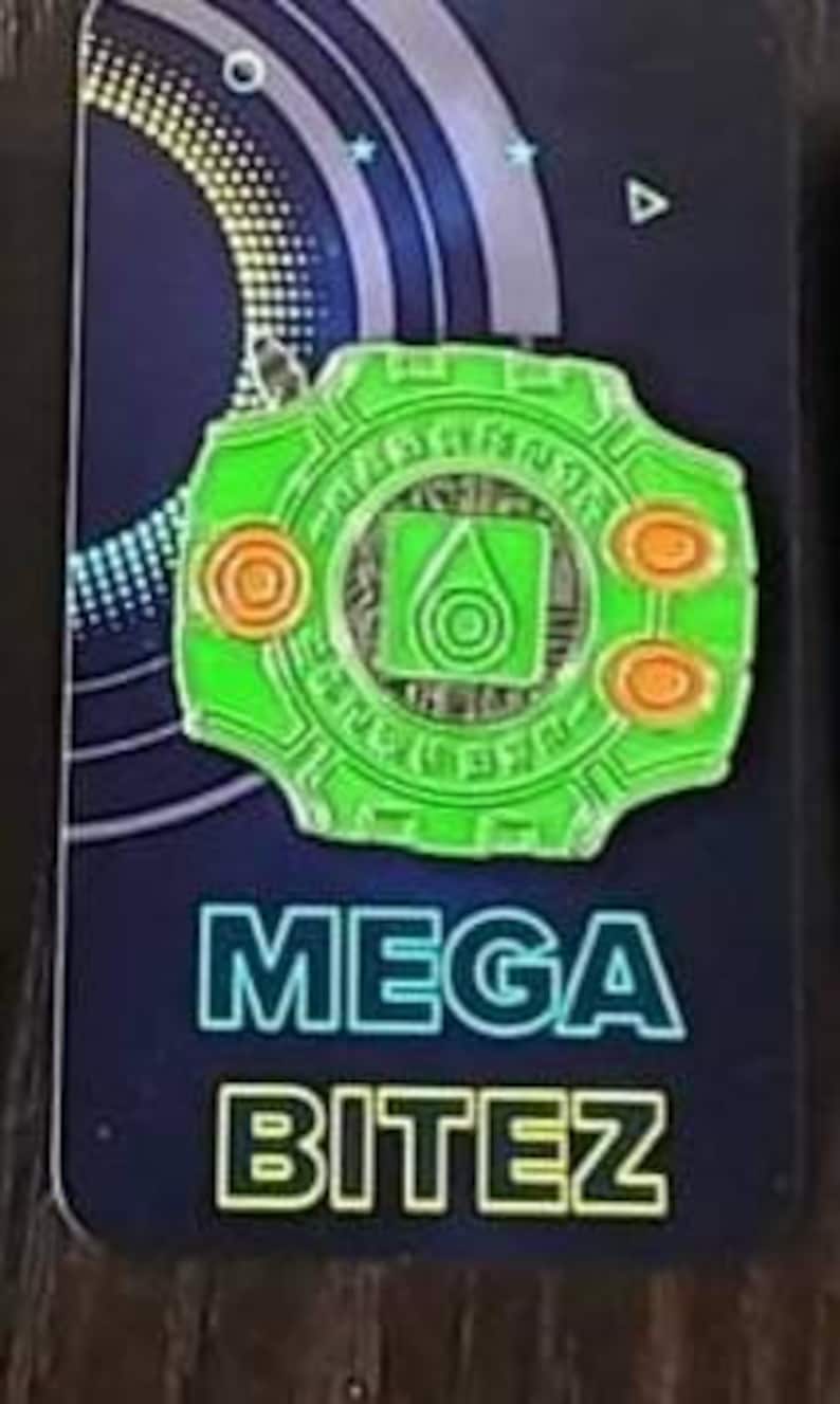 May include: Green and orange enamel pin featuring a stylized design of a digital device with the text "MEGA BITEZ" printed below.