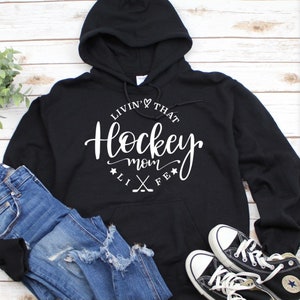 K&ouml;nnte beinhalten: Schwarzer Kapuzenpullover mit dem wei&szlig;en Schriftzug "Livin' That Hockey Mom Life". Das Design beinhaltet ein Herz, Sterne und gekreuzte Hockeyschl&auml;ger. Dazu blaue Jeans und schwarze Converse-Sneakers.