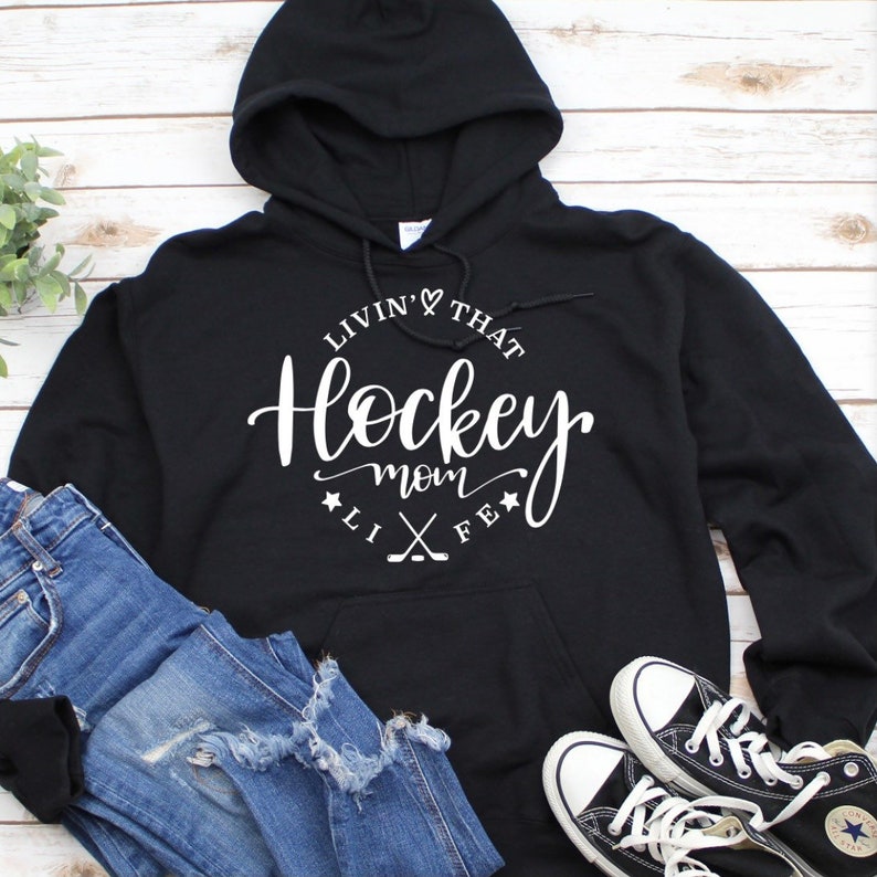 K&ouml;nnte beinhalten: Schwarzer Kapuzenpullover mit dem wei&szlig;en Schriftzug "Livin' That Hockey Mom Life". Das Design beinhaltet ein Herz, Sterne und gekreuzte Hockeyschl&auml;ger. Dazu blaue Jeans und schwarze Converse-Sneakers.