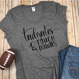 Pu&ograve; includere: Una t-shirt grigio antracite con scollo a V caratterizzata dalla scritta nera "tailgates & TOUCH DOWNS" stampata sul davanti. La maglietta &egrave; mostrata insieme a un pallone da football marrone e bianco e un paio di jeans blu su una superficie in legno chiaro.