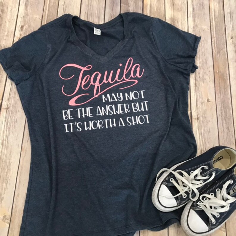 May include: Dark blue V-neck t-shirt with the text "Tequila may not be the answer but it's worth a shot" in pink and white. A pair of dark blue canvas sneakers with white laces and soles are next to the shirt.