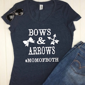 May include: A navy blue v-neck t-shirt with the words "BOWS & ARROWS" and the hashtag "#MOMOFBOTH" in white. The shirt is paired with blue jeans and sunglasses.