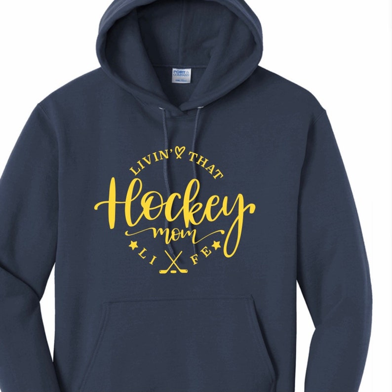 K&ouml;nnte beinhalten: Marineblauer Kapuzenpullover mit dem Text "Livin' That Hockey Mom Life" in gelber Schrift. Das Design umfasst ein Herz, Sterne und gekreuzte Hockeyschl&auml;ger. Der Pullover hat eine Vordertasche und eine Kapuze mit Kordelzug.