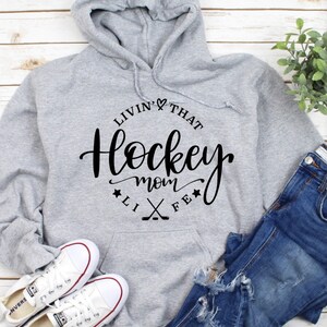K&ouml;nnte beinhalten: Grauer Kapuzenpullover mit dem schwarzen Schriftzug "Livin' That Hockey Mom Life". Das Design umfasst ein Herz, Sterne und gekreuzte Hockeyschl&auml;ger. Der Hoodie hat eine Kapuze mit Kordelzug und eine Vordertasche.
