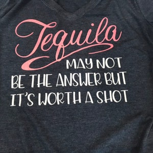 May include: Dark gray v-neck t-shirt with the text "Tequila may not be the answer but it's worth a shot" in pink and white lettering. The shirt is made of a soft material.